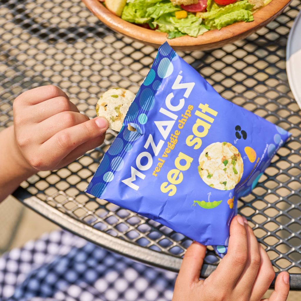 A blue single-serving bag of Mozaics Sea Salt Veggie Chips being held by a young person's hands outside at a patio table with a bowl of salad in the background.